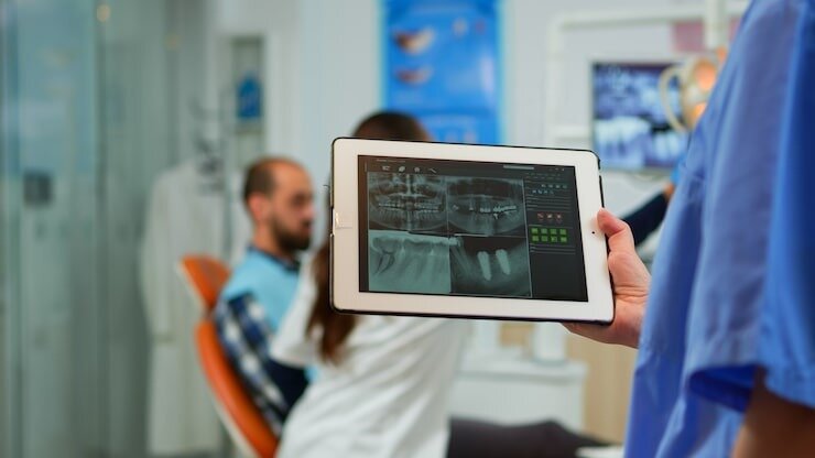 Close up dentist nurse holding tablet with digital radiography, while doctor is working with patient in background examining teeth problem sitting on stomatological chair in dental clinic.